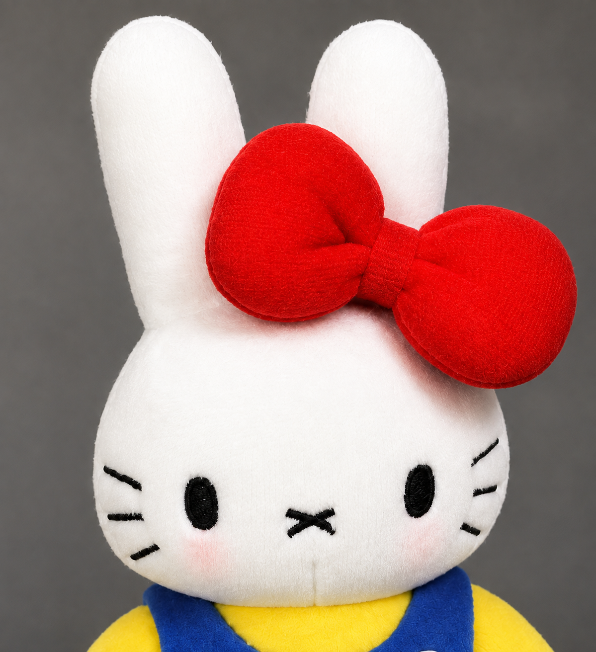 Close-up of white mini bunny plush with long ears, embroidered face, pink cheeks and oversized red bow.